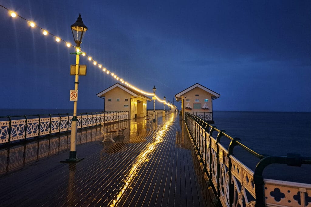 Mae goleuadau disglair ar linyn yn adlewyrchu oddi ar bier pren gwlyb yn y nos, gan arwain at ddau adeilad bach dros y môr. / Glowing string lights reflect off a wet wooden pier at night, leading to two small buildings over the ocean.